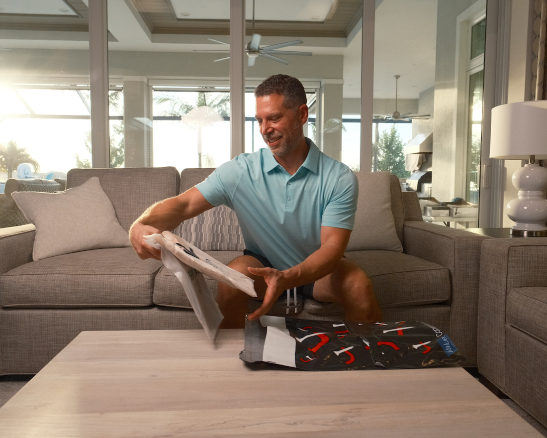 Man sitting on a couch in a living room holding a magazine and a bag of groceries.
