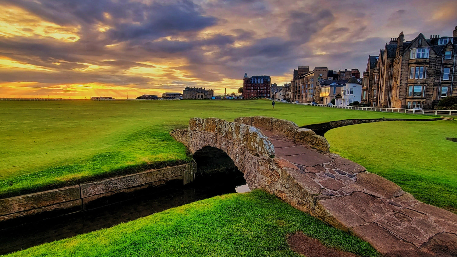 Golf course with a stone bridge and buildings under a colorful sky.